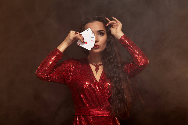 Young beautiful lucky brunette in a red evening dress shows her cards, a four of aces, hiding her face with cards - Фото, зображення