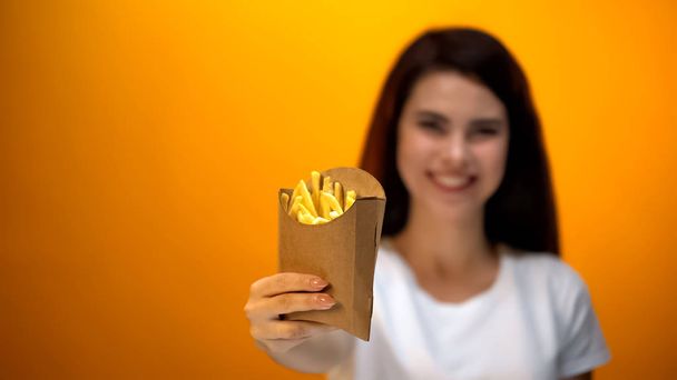 Sonriente chica mostrando papas fritas a la cámara, recomendando sabrosa comida rápida
 - Foto, Imagen