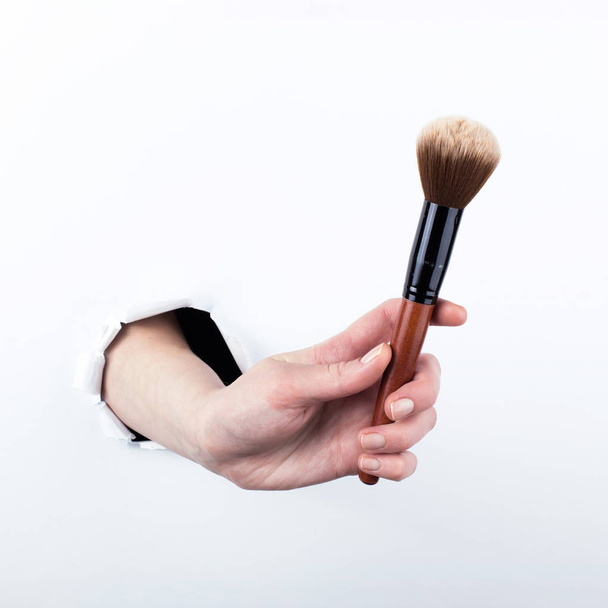 Female hand out of a hole in paper, holding a large powder brush. Isolate on white background. - Fotó, kép