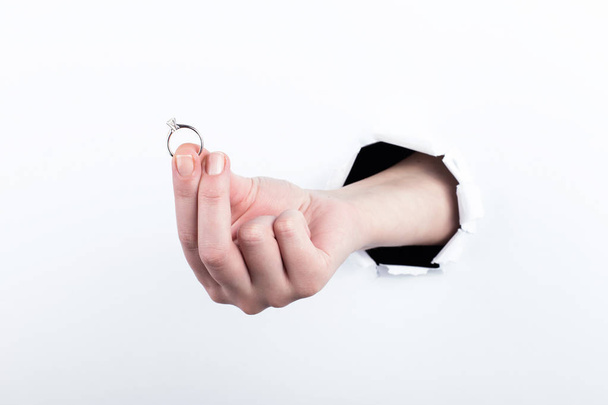 Female hand out of a hole in paper, holds a wedding ring, makes an engagement. Isolate on white background. - Foto, Bild