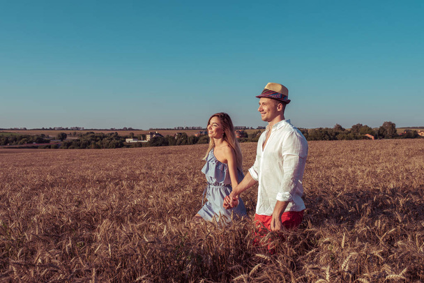 Beautiful young couple, man and woman, walking wheat field in summer, holding each others hand, smiling happy. Romantic walk in field, emotions love understanding. Support happy relationships. - Foto, Imagen