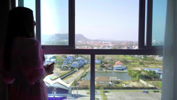 Nainen vaaleanpunainen läpinäkyvä aamutakki seisoo ja katsoo ulos panoraamaikkunaan ja juo kahvia. 4k Hua Hin, Thaimaa
. - Materiaali, video