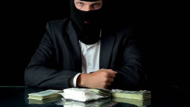 Mafiosi in balaclava looking at cocaine and money on table, drug trafficking - 写真・画像