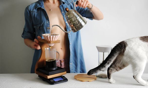 Красива дівчина в розтягнутій сорочці заварює каву з керамічною крапельницею орігамі. Білий кіт проходить повз. Безликий
 - Фото, зображення