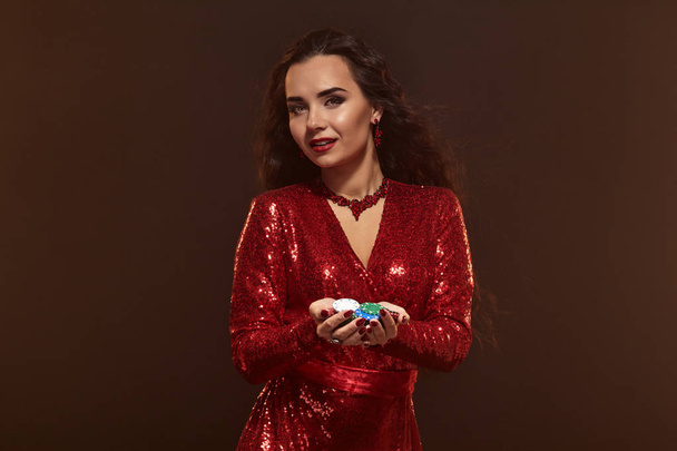 Young pretty lucky brunette woman in a red evening dress holds poker chips in her hands, showing her winnings. - Photo, Image