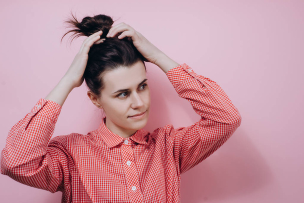 Horizontal portrait of young woman collecting hair in a ponytail, wearing red shirt , posing over pink background. Adorable female makes ponytail, advertises healthy natural hair - Valokuva, kuva