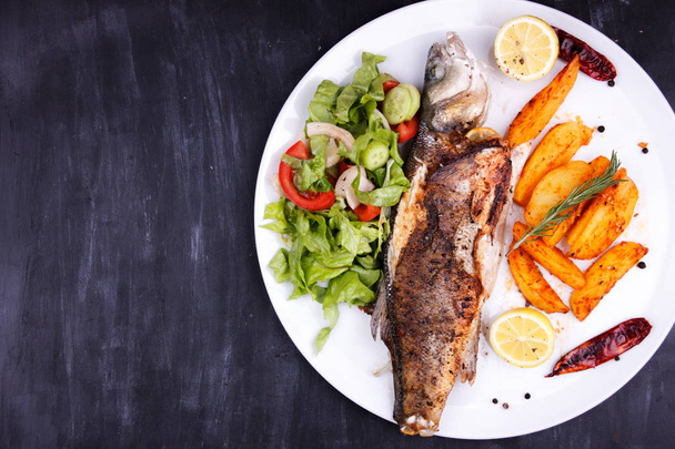 Fried sea bass with lemon, potatoes, salad and spices on a black wooden table - Photo, Image