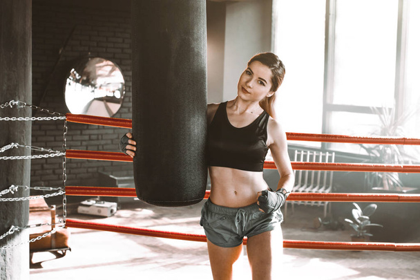 Female boxer training inside a boxing ring. Boxer practicing her moves at a boxing studio. The girl hugs a punching bag and looks at the camera - Zdjęcie, obraz