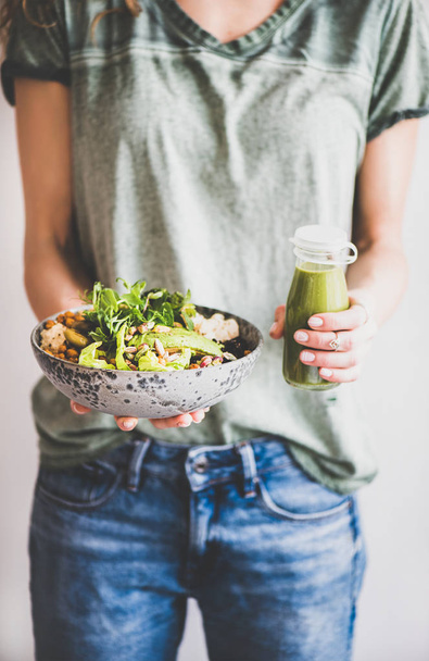 Healthy dinner or lunch. Woman in t-shirt and jeans standing and holding vegan superbowl or Buddha bowl with hummus, vegetable, fresh salad, beans, couscous and avocado and smoothie in hands - Фото, зображення