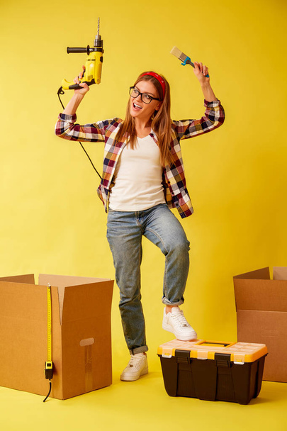 Woman skillfully holds a drill and a paint brush, standing between the boxes. Studio - Фото, изображение