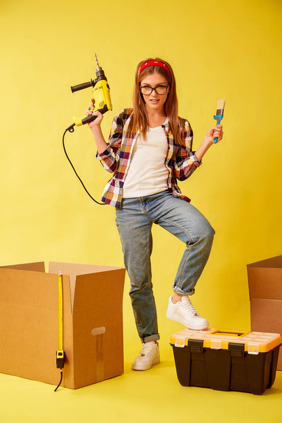 Woman skillfully holds a drill and a paint brush, standing between the boxes. Studio - Foto, immagini