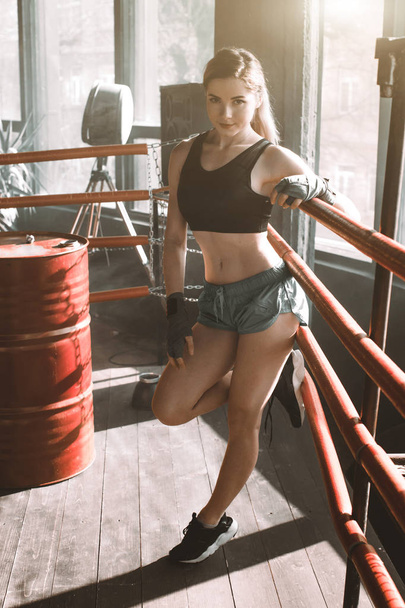 Young pretty boxer woman standing on ring. Full body portrait of boxer woman wearing black sports bra, grey trousers, trainers standing in ring and leaning on ropes. Boxing, sport concept - Zdjęcie, obraz