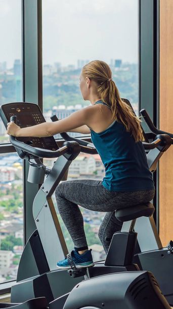 Young woman on a stationary bike in a gym on a big city background VERTICAL FORMAT for Instagram mobile story or stories size. Mobile wallpaper - Zdjęcie, obraz