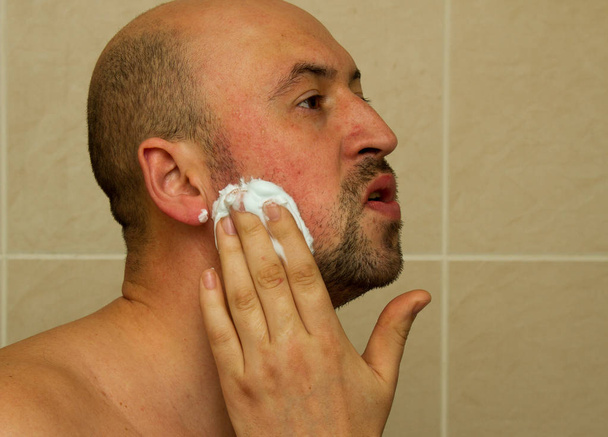 Portréja egy jó látszó fiatal férfi üzembe borotvakrém az arcán, és készen áll a borotválkozás a szakállát, a fürdőszobában - Fotó, kép