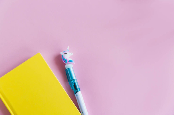 Caderno amarelo para notas, caneta unicórnio engraçado no fundo pastel rosa. Deitado. Vista superior. Espaço de cópia
 - Foto, Imagem