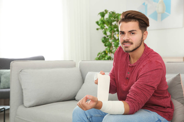 Young man applying bandage on injured arm at home, space for text. First aid - Φωτογραφία, εικόνα
