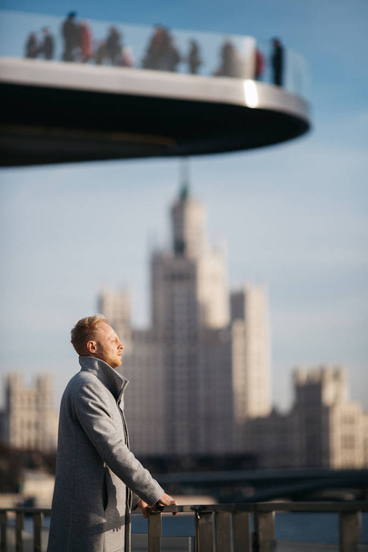 Photo of blond man in gray coat in city against background of building. - Zdjęcie, obraz