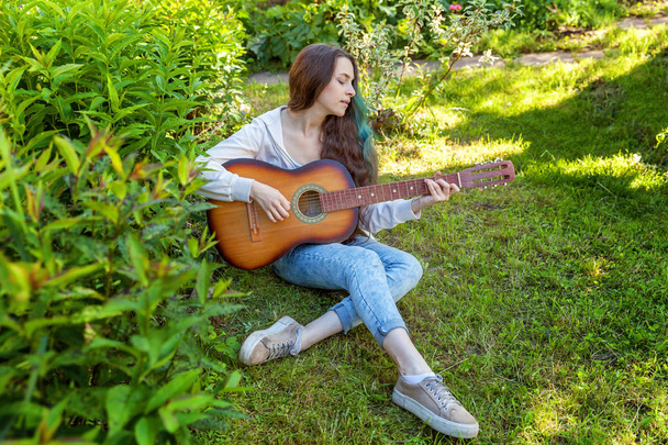 Young woman sitting in grass and playing guitar - Фото, изображение