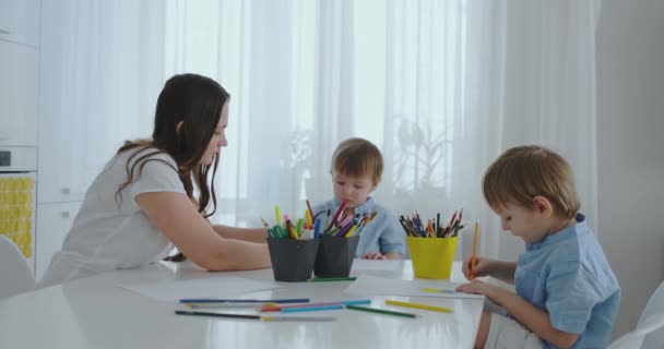 Mamá ayuda a los hijos a aprender a dibujar haciendo tareas de preparación preescolar en casa sentado en la cocina blanca
. - Imágenes, Vídeo