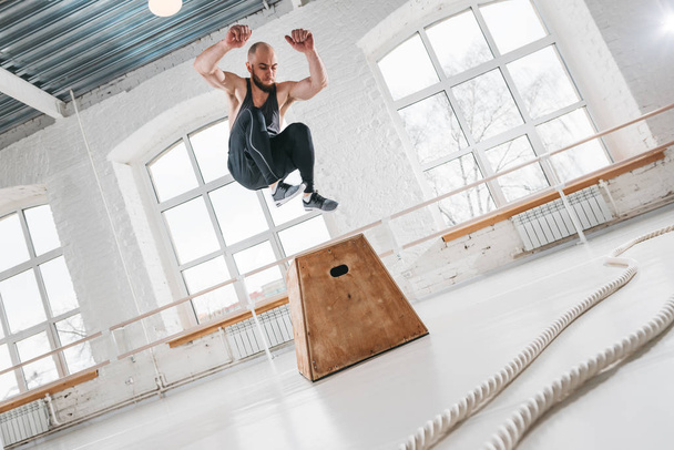 Dynamic shot of fitness male athlete jumping through at square box in crossfit gym. Strong man doing jump on wooden squat box at light hall - Фото, изображение