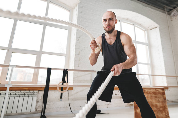 Strong man workout with battle ropes at light gym. Muscular sportsman doing crossfit excursion with ropes in workout gym - Photo, image