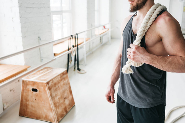 Handsome muscular man holding on shoulder rope workout in light gym. Portrait of fitness man with battle ropes at cross gym - 写真・画像