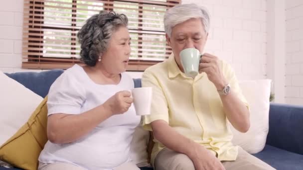 Asian elderly couple drinking warm coffee and talking together in living room at home, couple enjoy love moment while lying on sofa when relaxed at home. Lifestyle senior family at home concept. - Footage, Video