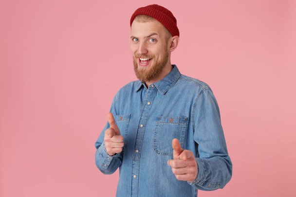 Bearded male in fashioned denim shirt red hat, encourages,smiles, makes supportive gesture, pointing on camera as on chosen, best player, isolated over pink background. Human body language - Photo, Image