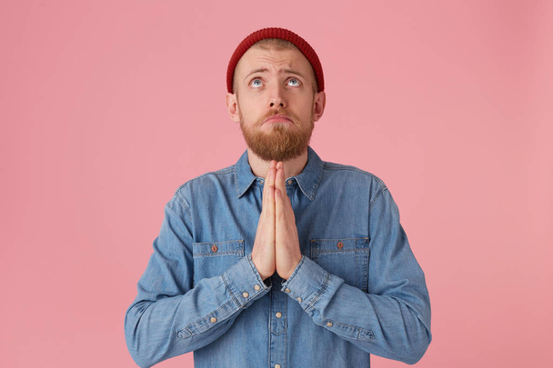 Young bearded guy looks up with hope, palms folded in a prayer gesture, asking for help from higher powers, protection, blessing, wearing a denim shirt, isolated on a pink background - Photo, Image