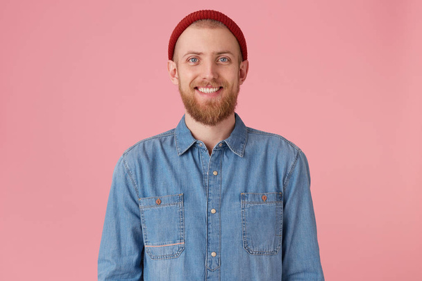 Smiling blue-eyed guy in red hat with red thick beard feels happiness joy gladness showing white healthy teeth, wears fashionable denim shirt, isolated on pink background - Фото, зображення