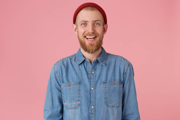 Laughing joyful guy in red hat with red thick beard feels happiness joy gladness showing white healthy teeth, wears fashionable denim shirt, isolated on pink background - Foto, Bild
