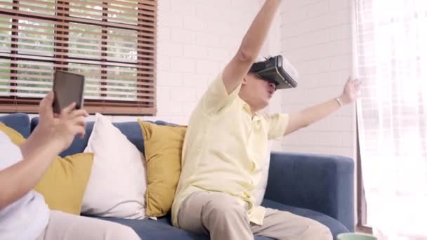 Asian elderly couple using tablet and virtual reality simulator playing games in living room, couple feeling happy using time together lying on sofa at home. Lifestyle Senior family at home concept. - Filmagem, Vídeo
