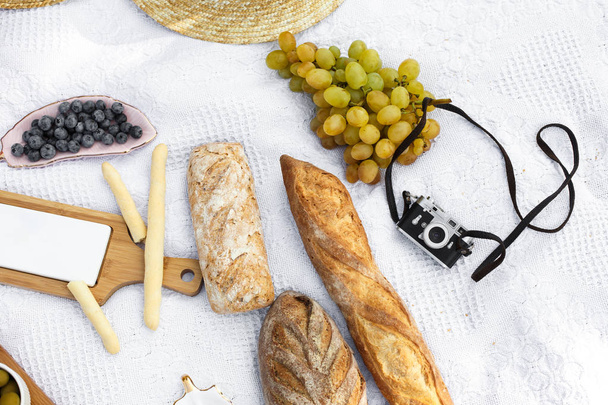 Food lay out on picnic blanket. fresh baked bread, grapes, olives and photocam lay on white blanket. Picnic prepare decoration - Photo, image