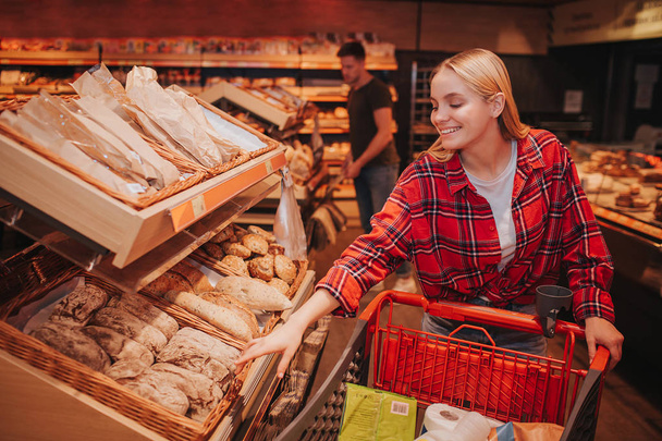 Young couple in grocery store. Woman reach to bread with hand and smile. Guy stand behind and pick food. - Photo, Image
