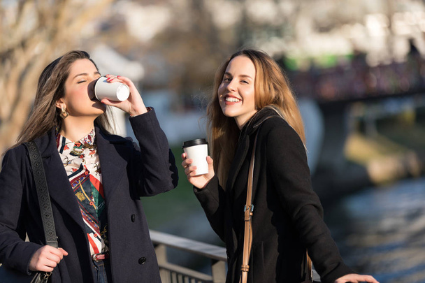 Buiten mode portret van twee jonge mooie vrouwen vrienden koffie drinken. Glimlachend en gaan winkelen. Zoenen een kopje koffie. Heldere make-up - Foto, afbeelding