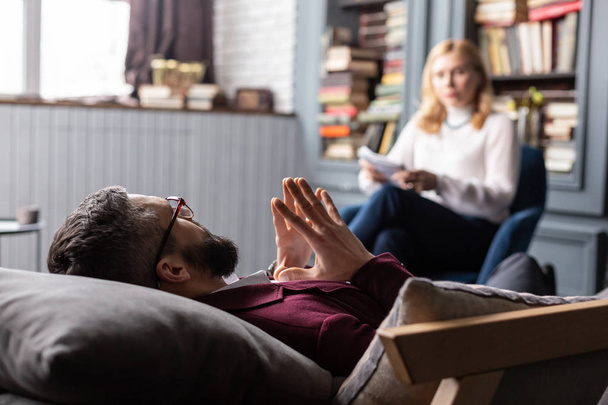 Bearded man in glasses lying on sofa while talking to therapist - Photo, Image