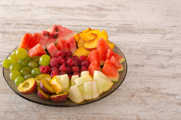 Μείγμα φρούτων. Ροδάκινο, σταφύλια, μήλο, καρπούζι, πεπόνι, βατόμουρο, δαμάσκηνο. Κομμάτια φρούτων σε ένα πιάτο βρίσκονται σε λευκό φόντο ξύλινη. Αντιγράψτε το χώρο, χώρο για το κείμενο. Επίπεδη Lay - Φωτογραφία, εικόνα