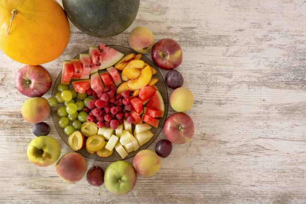 Hedelmäsekoitus. Omena, persikka, rypäleet, vesimeloni, meloni, vadelma, luumu. Kokonaiset hedelmät ja viipaleet lautasella ovat valkoisella puupohjalla. Kopioi tilaa, Tekstipaikka. Tasainen makuuasento
 - Valokuva, kuva