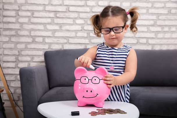 Ευτυχισμένο χαριτωμένο κορίτσι ζωγραφική για Piggy Τράπεζα με μαύρο μαρκαδόρο στο σπίτι - Φωτογραφία, εικόνα