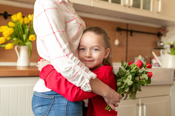 Happy Mother's Day or Birthday Background. Adorable young girl hugging her mom after surprising her with bouquet of red roses. Family celebration concept. - Photo, image