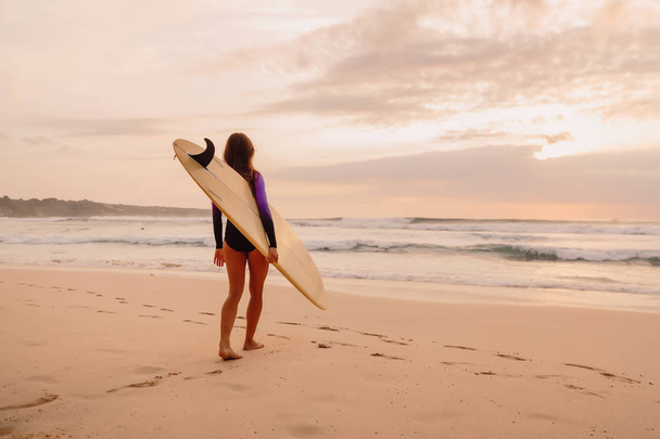 Surfer girl with surfboard on a beach at sunset or sunrise. - Фото, изображение