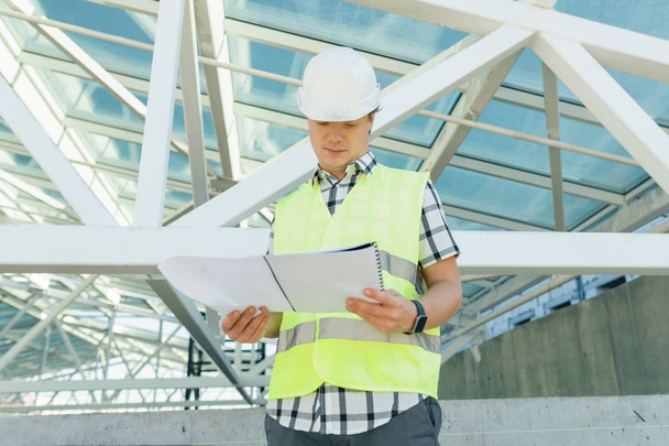 Portrait of male engineer at construction site, builder looks at blueprint. Building, development, teamwork and people concept - Φωτογραφία, εικόνα