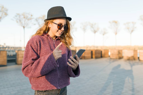 Young girl teenager in hat and glasses looking in mobile phone and showing index finger up attention, idea, eureka, golden hour. - Фото, зображення