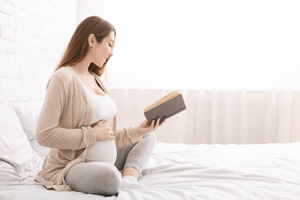Smiling pregnancy reading ibook sitting on bed - Valokuva, kuva