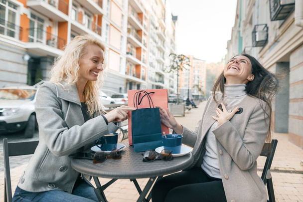 Two young beautiful women in an outdoor cafe with shopping bags and cup of coffee. Enjoying women looking at purchases - 写真・画像