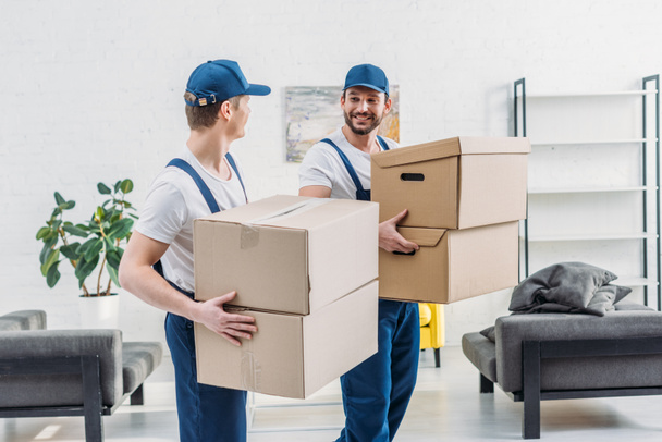 two movers in uniform looking at each other while carrying cardboard boxes in apartment - Photo, Image
