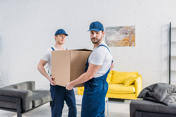 two handsome movers in uniform transporting cardboard box in apartment - Foto, Imagen