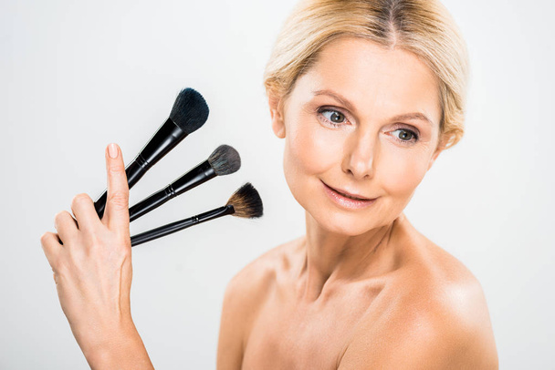 beautiful and blonde mature woman looking away and holding cosmetic brushes on grey background  - Фото, изображение