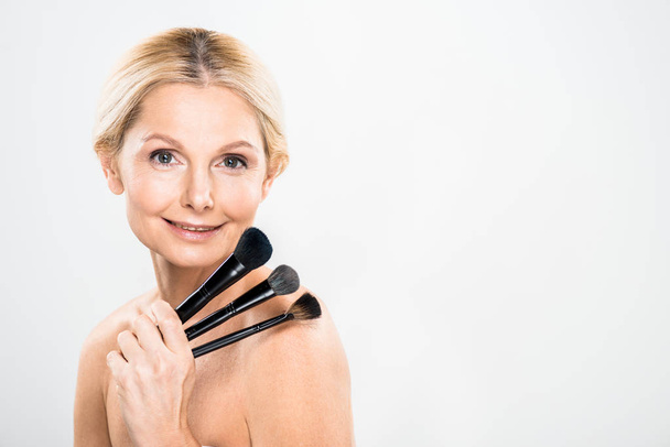 beautiful and blonde mature woman looking at camera and holding cosmetic brushes on grey background  - Foto, Imagem