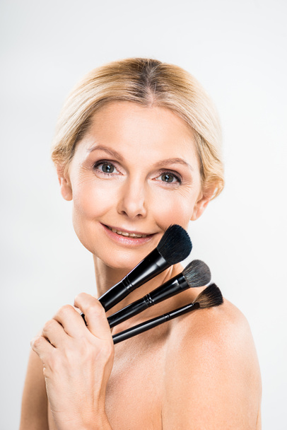beautiful and smiling mature woman looking at camera and holding cosmetic brushes on grey background  - Photo, Image